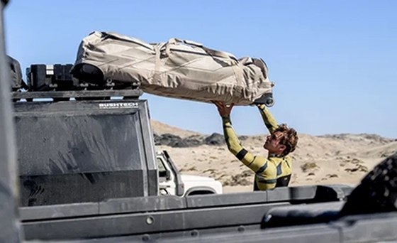 A kiter lifting a packed boardbag onto the roof rack of a pickup truck in the desert