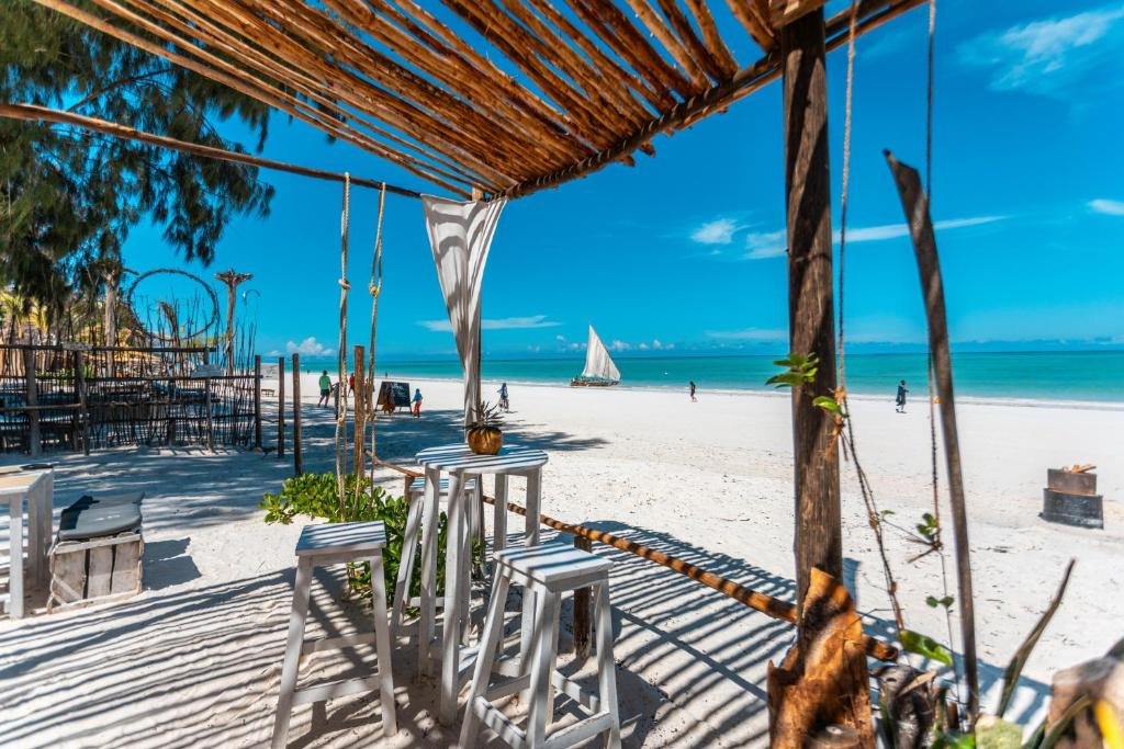White wooden stools under a reed roof on Paje beach, with a dhow sailing on the turquoise lagoon